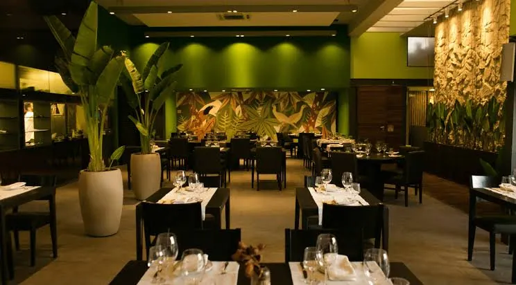 Salão do restaurante com mesas postas e painel de folhagens ao fundo